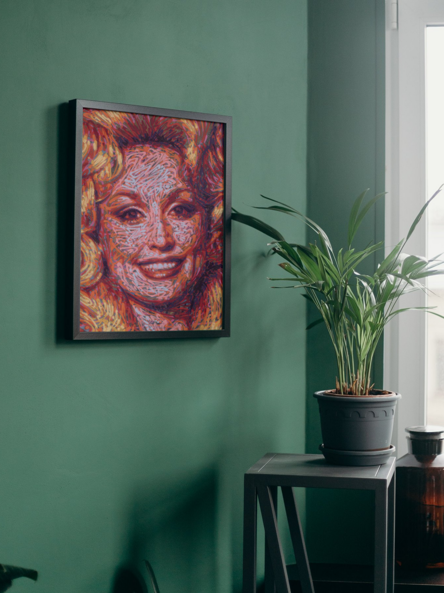 Mockup of a graffiti spray can portrait by Ben Jay of Blank Space Removal of a closeup of Dolly Parton's face and bouffant hair, in a black frame hung on a green wall with a houseplant nearby.