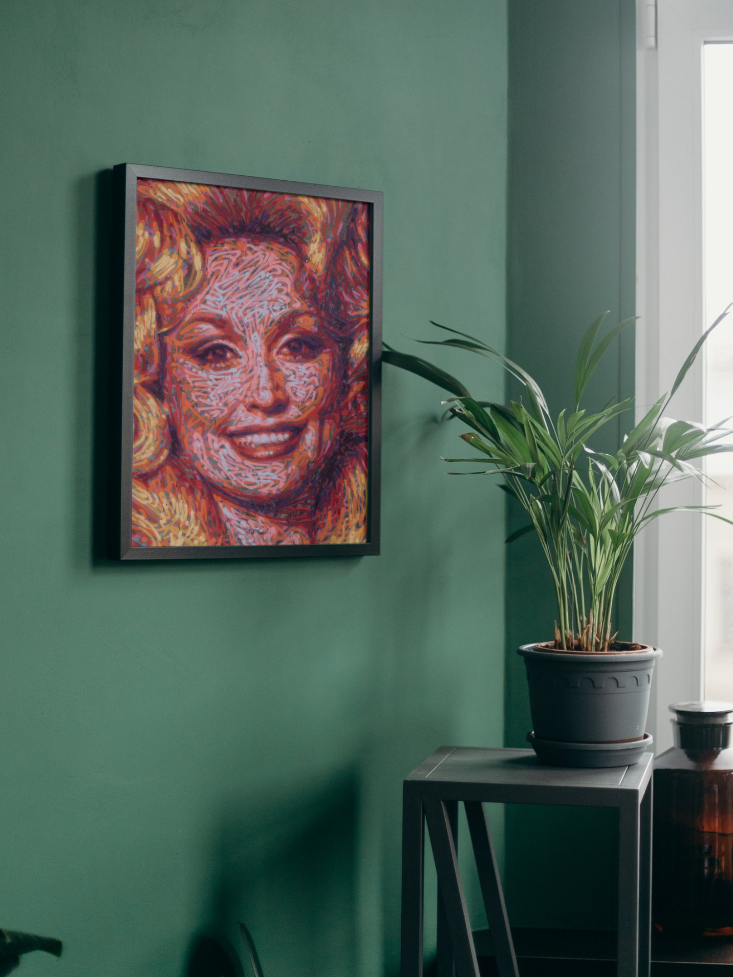 Mockup of a graffiti spray can portrait by Ben Jay of Blank Space Removal of a closeup of Dolly Parton's face and bouffant hair, in a black frame hung on a green wall with a houseplant nearby.