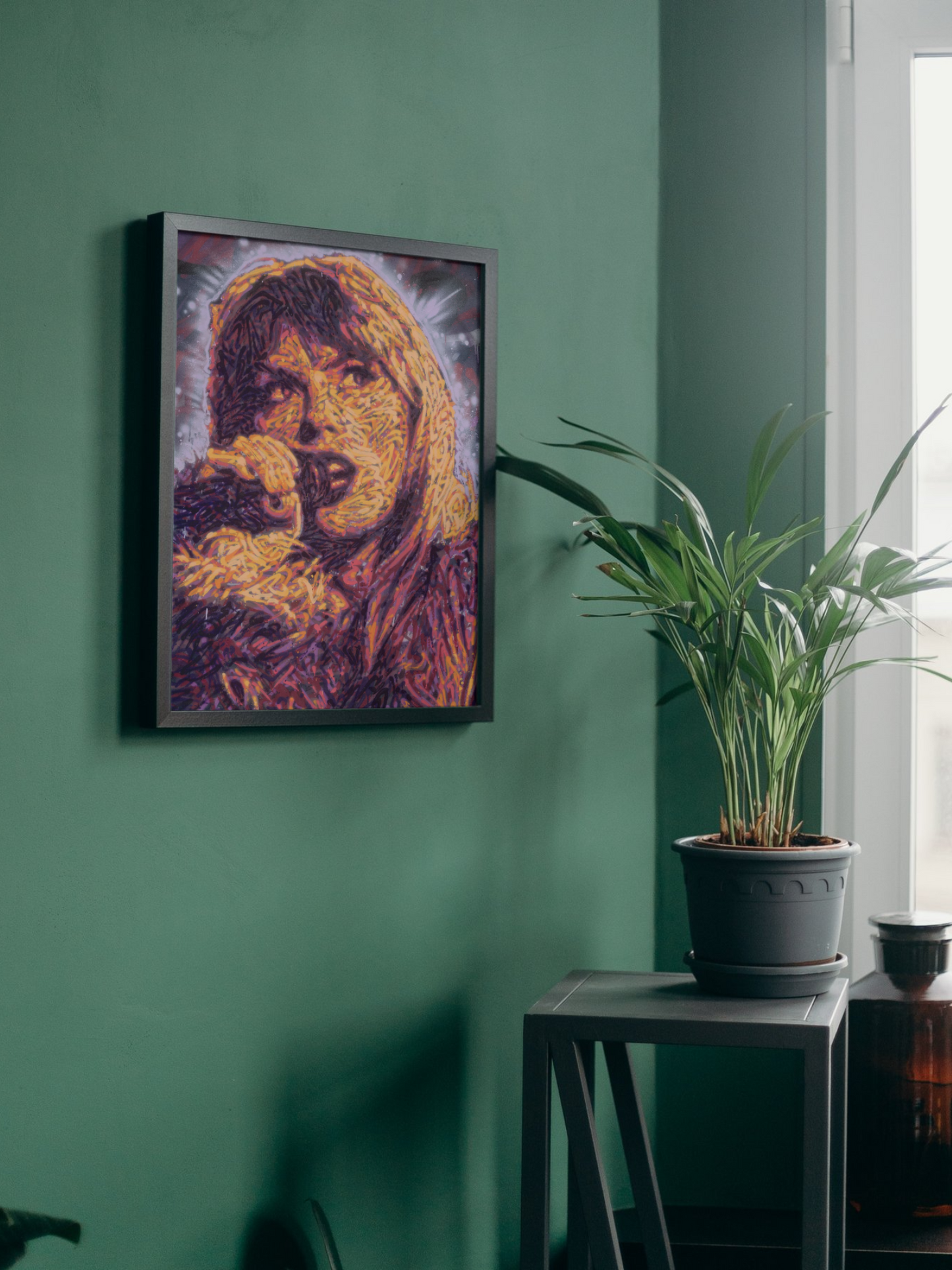 Mockup of a graffiti spray can portrait by Ben Jay of Blank Space Removal of Taylor Swift holding a microphone, in a black frame hung on a green wall with a houseplant nearby.